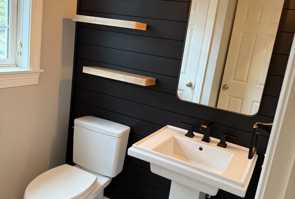 A modern bathroom featuring a black shiplap wall, a white pedestal sink, a toilet, and two wooden shelves illuminated by stylish wall sconces