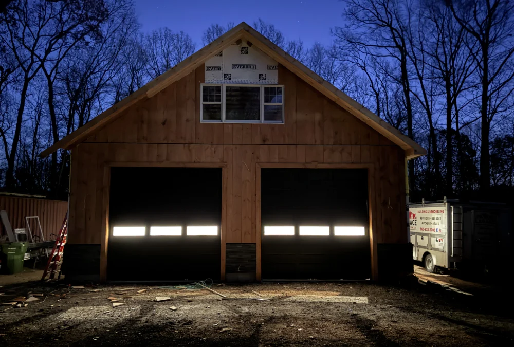 A newly constructed wooden garage at dusk, featuring illuminated garage doors and surrounding trees silhouetted against a dark blue sky.