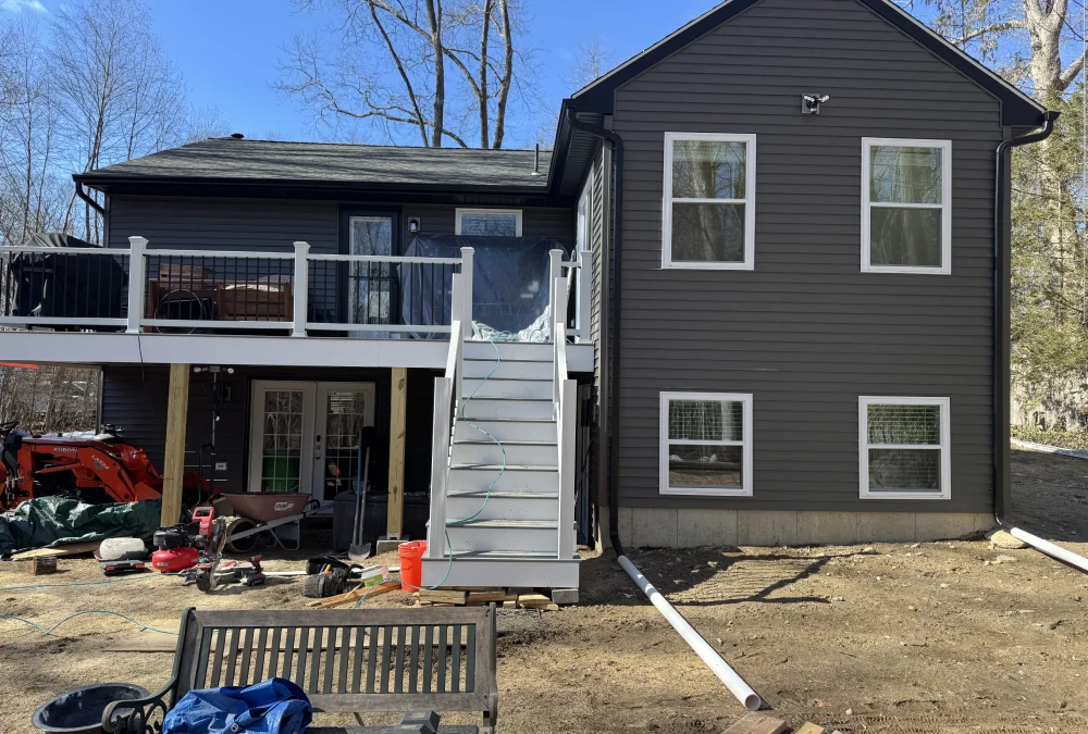 Rear view of a two-story gray house with a deck, stairs, and construction materials scattered in the yard under a clear blue sky.