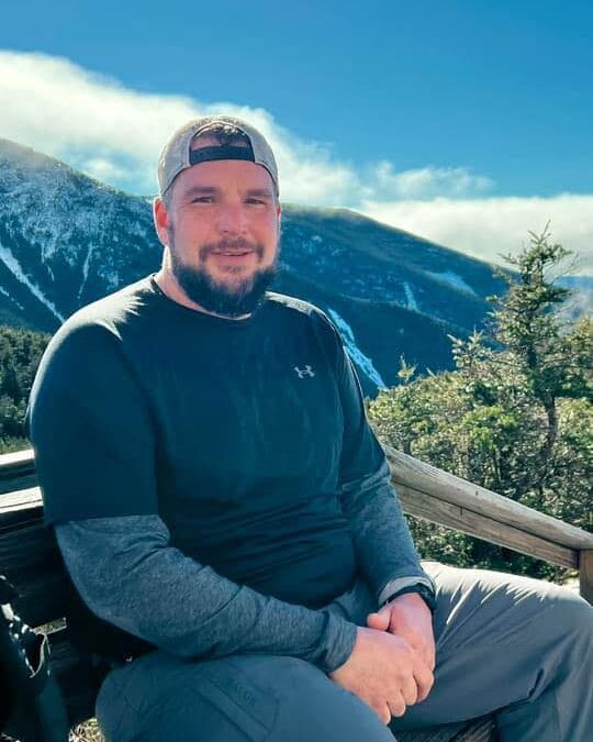 Scott Lagace sitting on a wooden bench with mountains and blue sky in the background, enjoying a scenic outdoor view