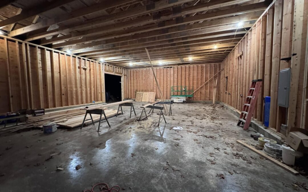 Large unfinished interior space with exposed wooden framing and open ceiling joists, prepared for remodeling. The area features a concrete floor, scattered construction tools, sawhorses, a red ladder, and stacked lumber. Overhead work lights illuminate the structure. Located in Columbia, Connecticut.