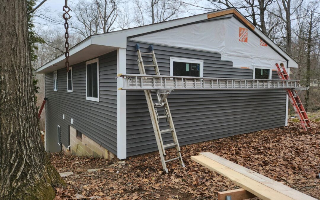 House on a sloped lot undergoing exterior renovation with new dark gray vinyl siding being installed. The upper portion is still wrapped in Home Depot-branded house wrap. Several ladders and construction tools are positioned around the site, with fallen leaves covering the ground. Located in Andover, Connecticut.